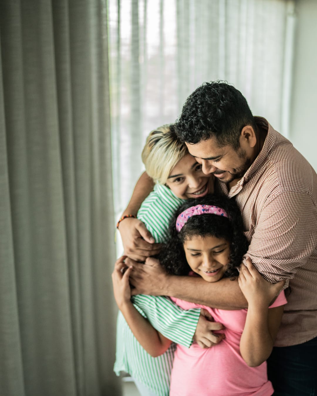 A father, mother and daughter hugging indoors beside a window with sheer curtains, all smiling